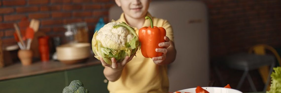 Little child demonstrating fresh red pepper and cauliflower and looking at camera while standing near table with vegetables and ingredients put into deep ceramic salad bowl in kitchen