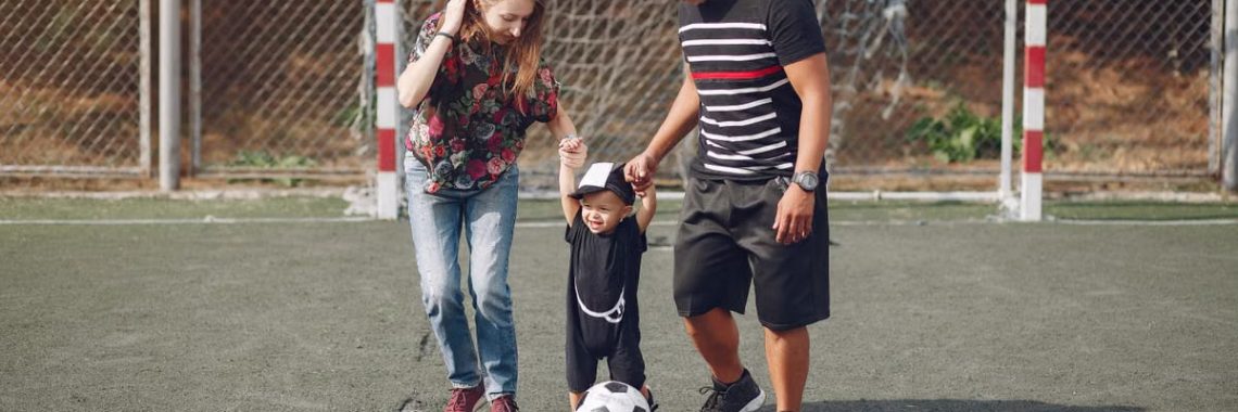 Full body of positive happy parents in casual clothes holding toddler son hands while walking together on football field on sunny day